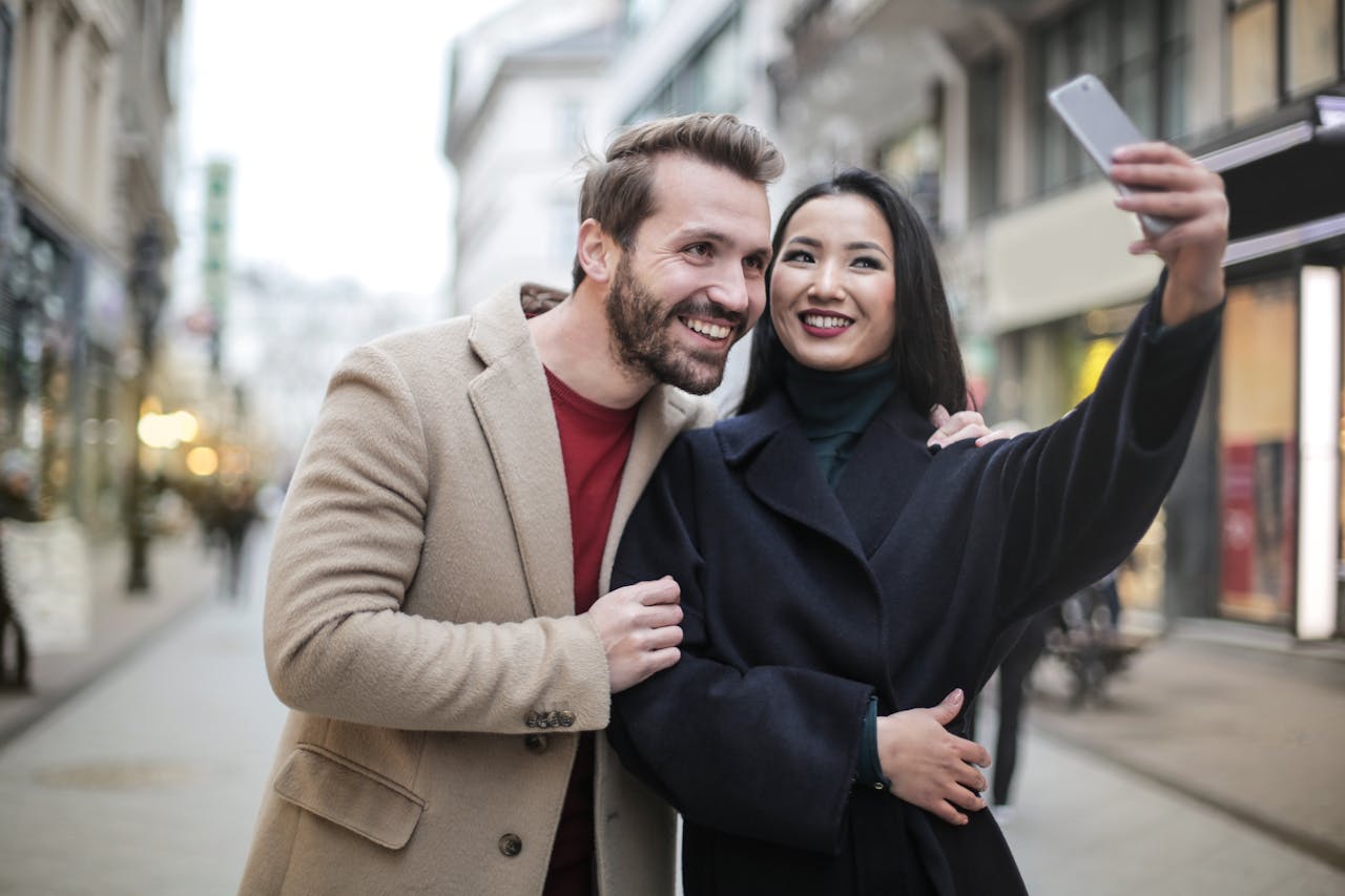 Features Young couple smiling and taking a selfie in a city street during daytime.