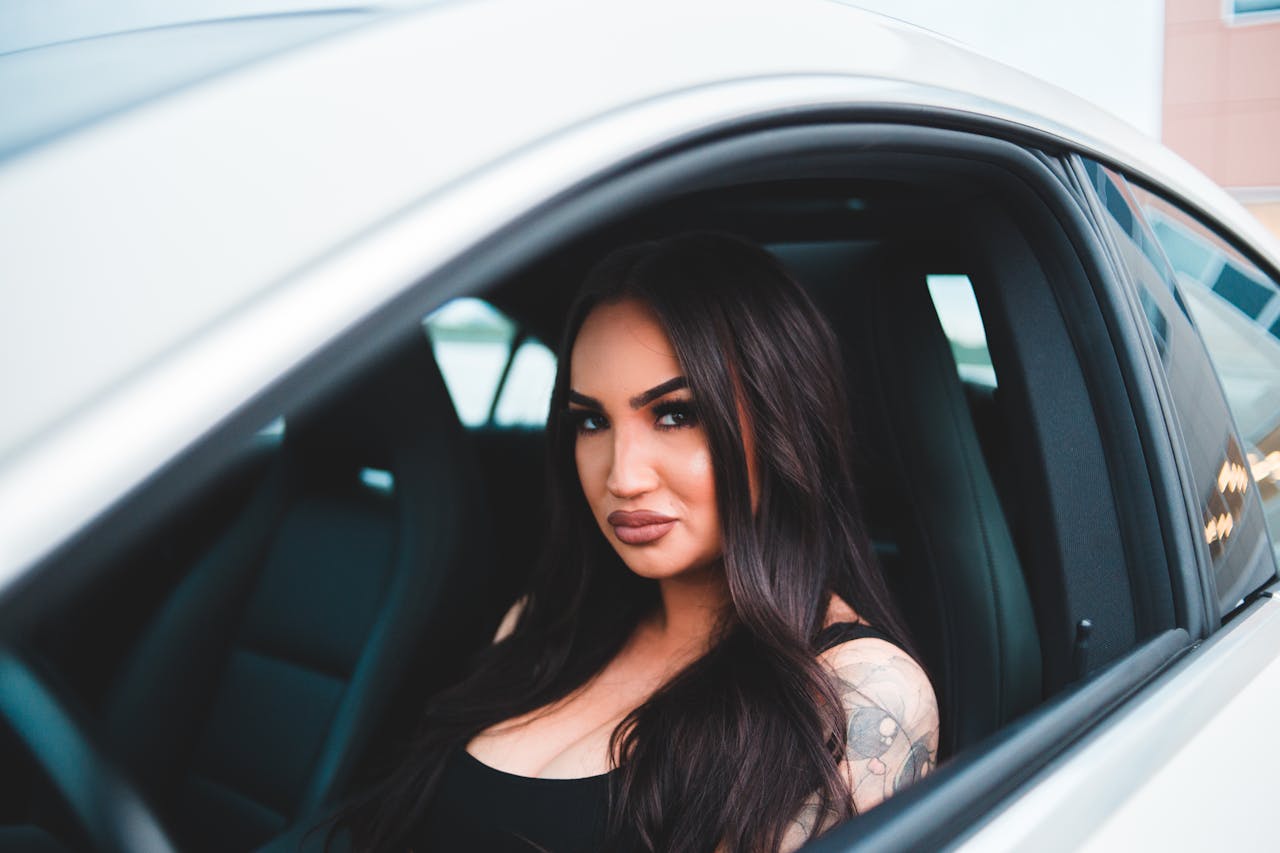Home Gorgeous female with long dark hair and bright makeup looking at camera while sitting in front of steering wheel in cabin of automobile